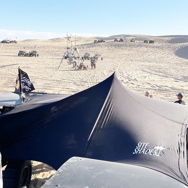 Black shade sail on a vehicle with 'SITE SHADES' branding, set against a desert landscape with people and vehicles in the background.